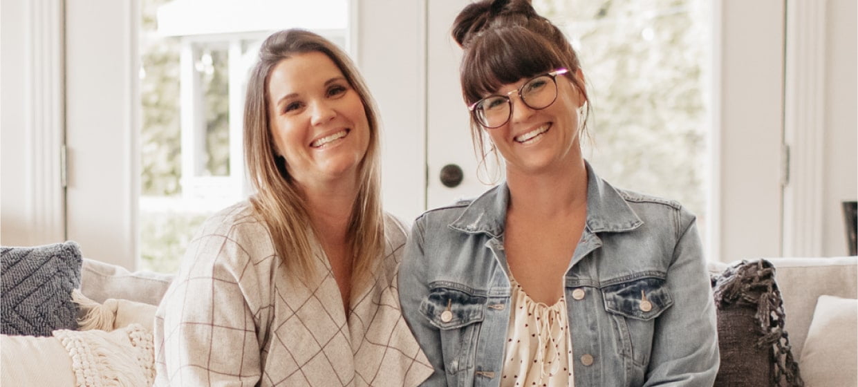 Lyndsay and Leslie from Lamb & Co and HGTV's Unsellable Houses, looking at the camera and smiling while sitting next to each other on a couch