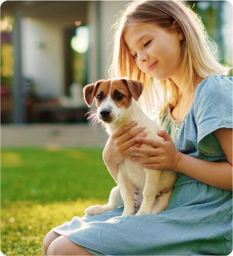 A young girl in a blue dress sitting on her knees and holding a small dog in a backyard